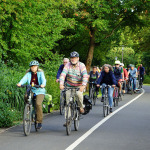 Teilnehmer einer mobilen Bürgerversammlung in Nürnberg auf ihren Fahrrädern. Sie fahren auf einem Radweg durch eine grünbewachsene Umgebung.