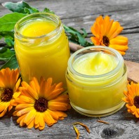 Zwei Gläschen mit selbstgemachter Calendulacreme. Daneben liegen zwei orangefarbenen Calendulablüten auf einem Holztisch.